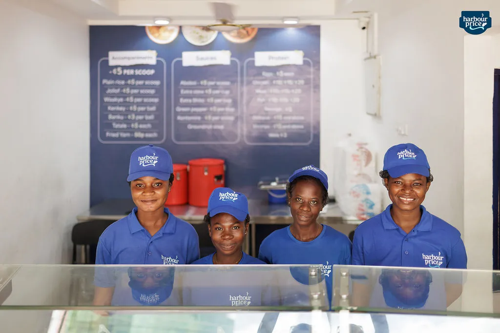 Hot Meals servers in royal blue caps smiling behind the glass food counter
