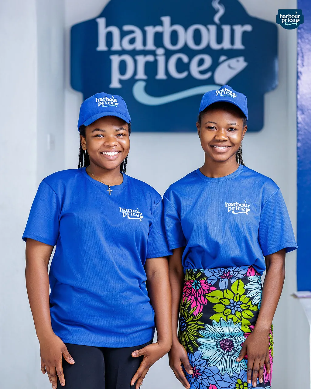 Two smiling Harbour Price staff in royal blue caps and tees under the big Harbour Price sign