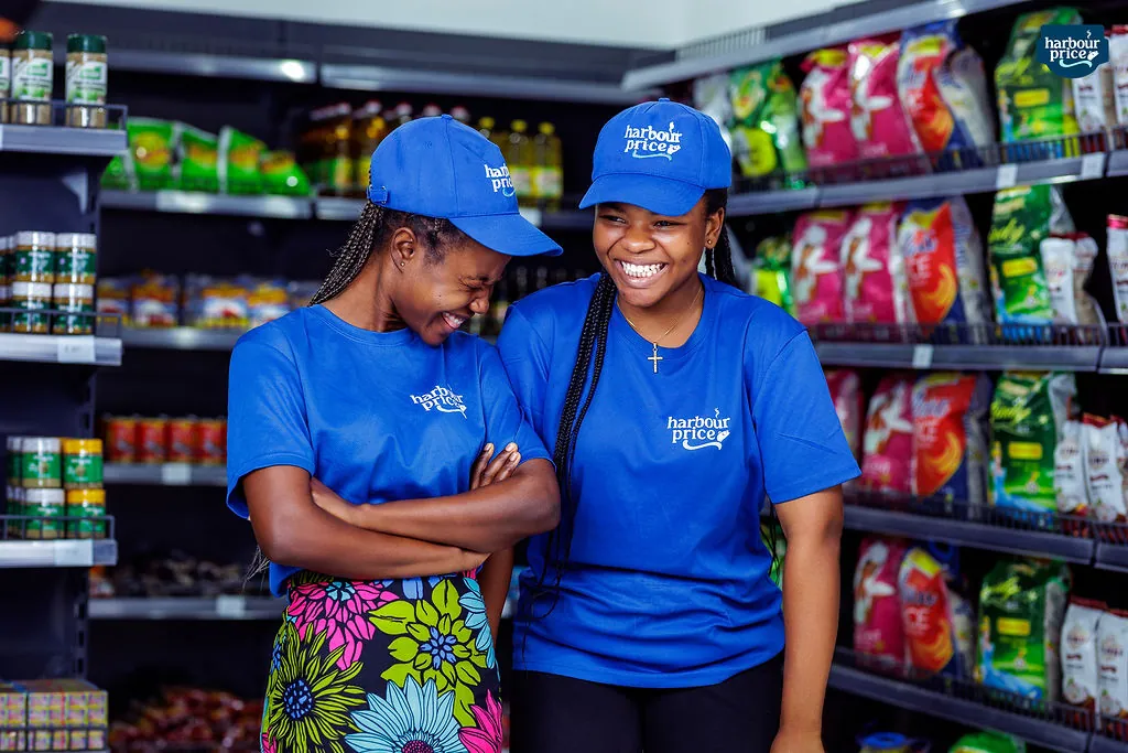 Two Harbour Price staff laughing together in a grocery aisle, arms folded